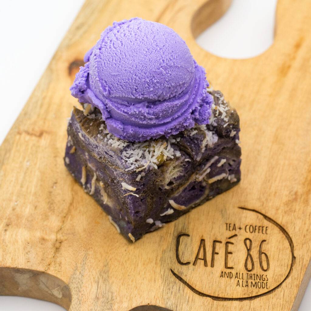 A square ube brownie with shredded coconut topped with a scoop of purple ube ice cream, served on a wooden Café 86 board.